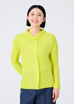 A smiling person with straight black hair wears the KNIT STRETCH PLEATS 1 shirt by me ISSEY MIYAKE in bright yellow and blue pants, standing comfortably against a plain white background.