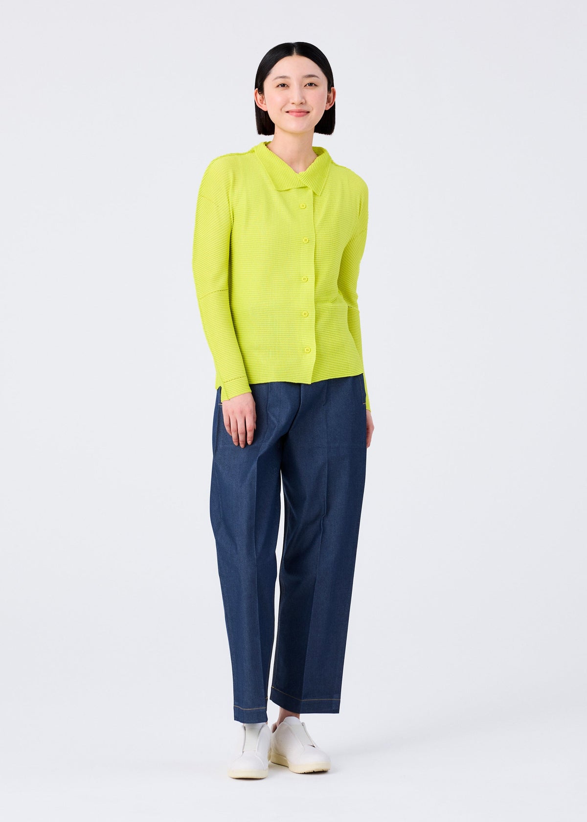 A woman smiles against a white background, wearing the me ISSEY MIYAKE KNIT STRETCH PLEATS 1 shirt in bright yellow-green and loose blue pants with subtle stretch pleats, paired with white shoes. view 8 of 8