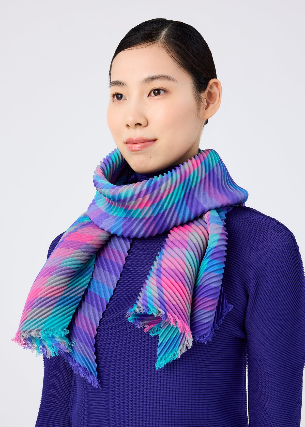 A woman with straight dark hair wears the CHECK PLEATS STOLE from me ISSEY MIYAKE—a vibrant, yarn-dyed piece with blue, purple, pink, and turquoise stripes—draped over a textured deep purple top against a light background. view 6 of 6