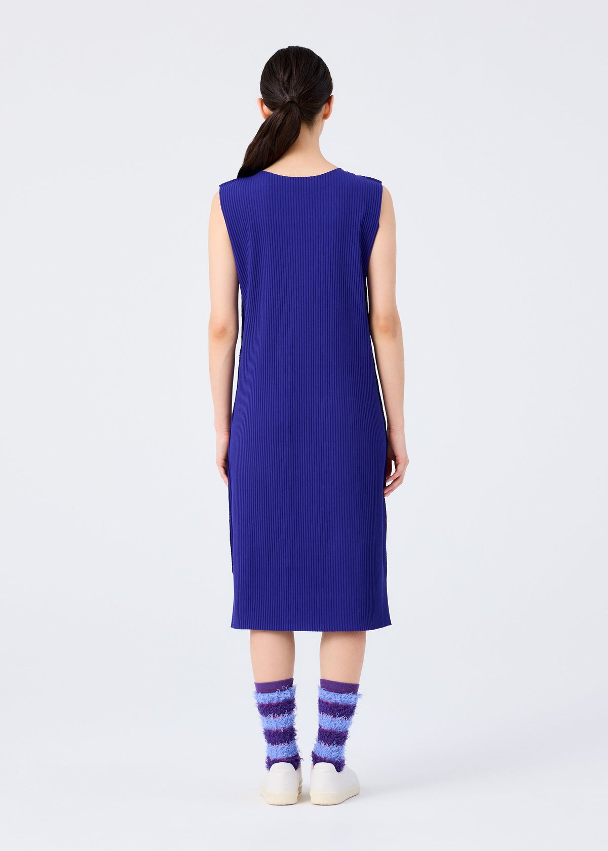 A woman with long dark hair in a low ponytail stands with her back to the camera, wearing a blue dress and me ISSEY MIYAKE TAIL SOCKS, paired with white shoes, against a plain light background. view 7 of 7