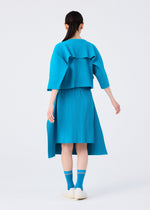 A person with long dark hair, dressed in a bright blue, pleated outfit, stands facing away against a plain white background, wearing CARD SOCKS by me ISSEY MIYAKE and white shoes.