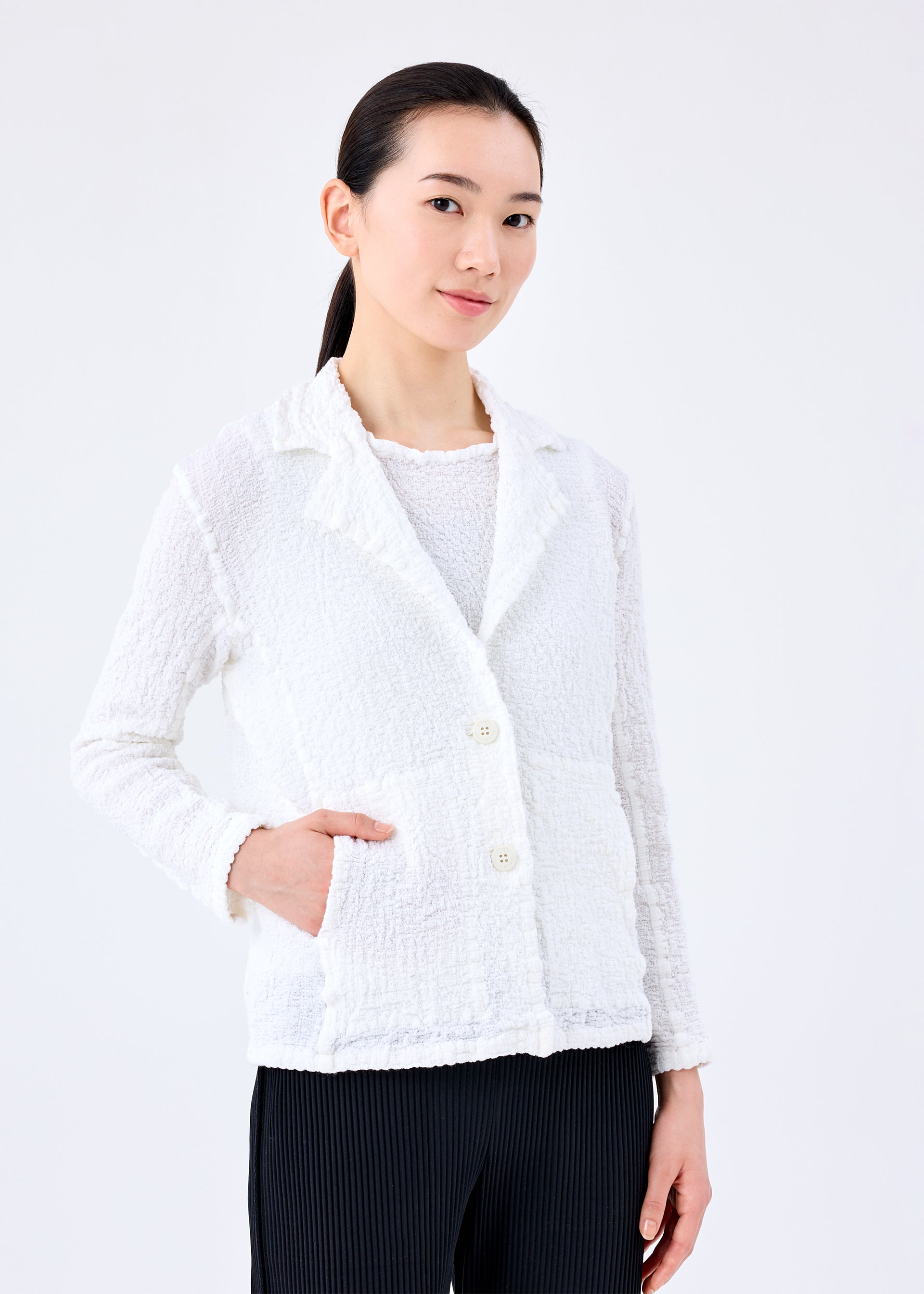 Zoomed view A woman with dark hair in a low ponytail wears the me ISSEY MIYAKE CAULIFLOWER JACKET over a white top and black pants, standing against a plain white background with one hand in her pocket and a slight smile.