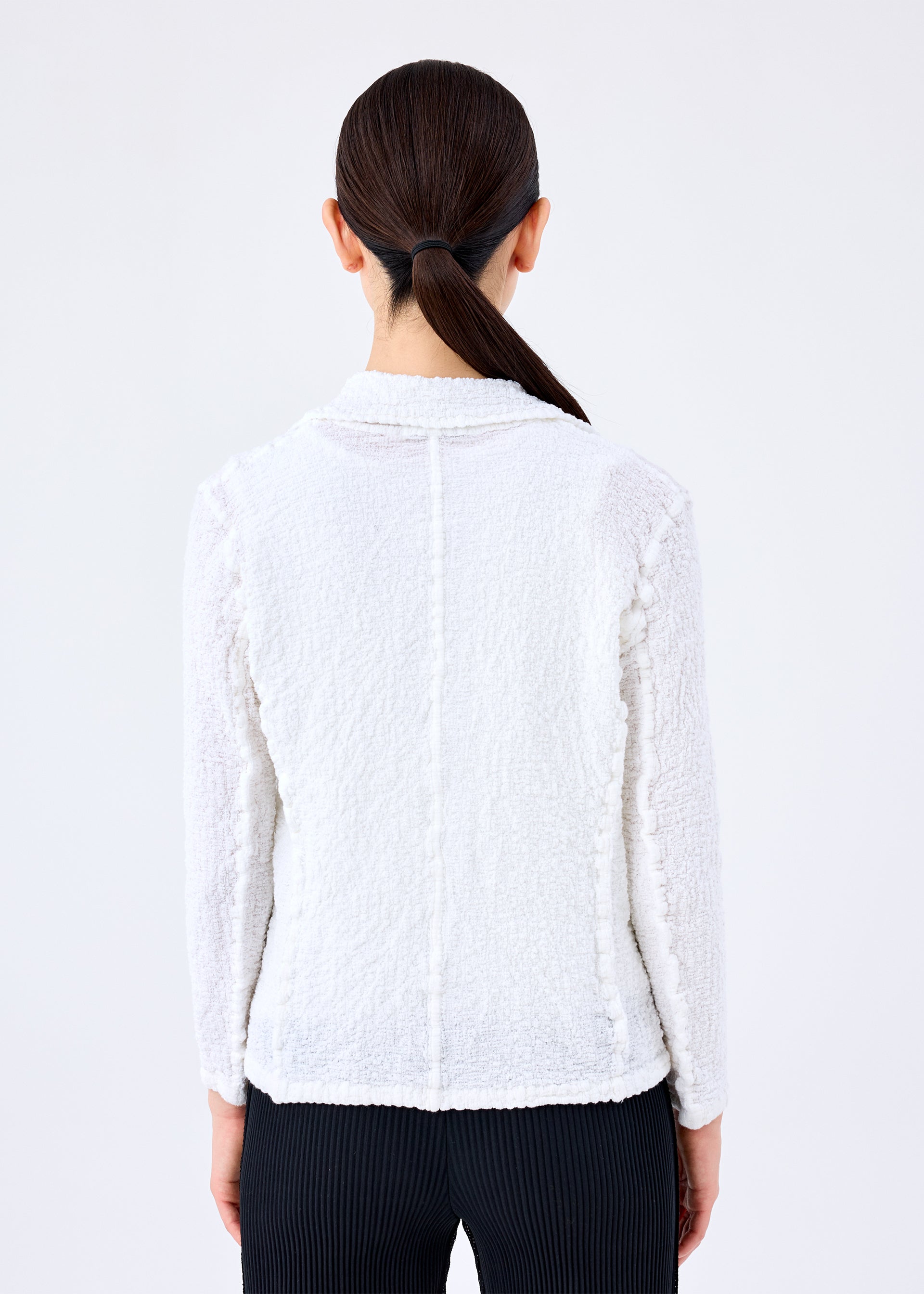 Zoomed view A person with straight dark hair in a low ponytail is shown from behind, wearing the white textured CAULIFLOWER JACKET by me ISSEY MIYAKE and black pants against a plain light background.