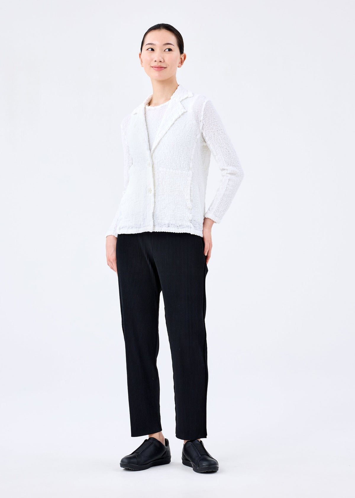 A person stands against a plain white background, wearing the CAULIFLOWER JACKET by me ISSEY MIYAKE in textured fabric, paired with a white top, black pinstripe pants, and black shoes. They look slightly to the side and smile softly. view 6 of 6