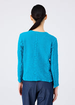 A person with long dark hair in a low ponytail is seen from behind, wearing the textured bright blue CAULIFLOWER COLOR jacket and dark blue pants by me ISSEY MIYAKE against a plain light background.