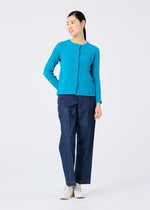 A woman wearing the bright blue "CAULIFLOWER COLOR" jacket by me ISSEY MIYAKE stands against a white background, paired with dark blue pants and white shoes. She smiles slightly, looking to the side with one hand on her hip.
