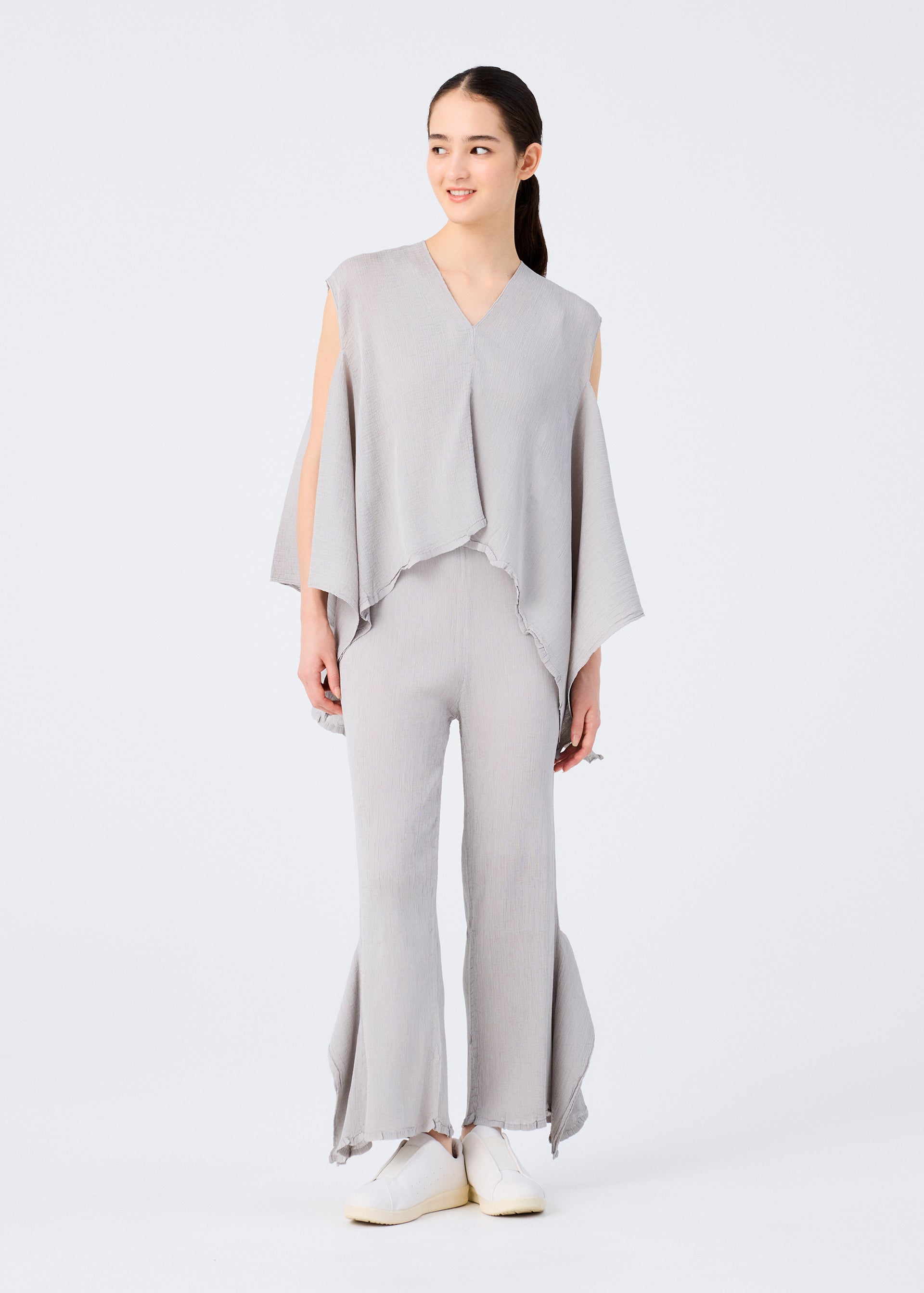 Zoomed view A woman models the me ISSEY MIYAKE FLAT SEE-THROUGH CREPE set: a light gray, loose-fit poncho top with open sleeves and matching flared pants with frayed hems. She pairs white shoes and has her hair tied back against a plain white background.