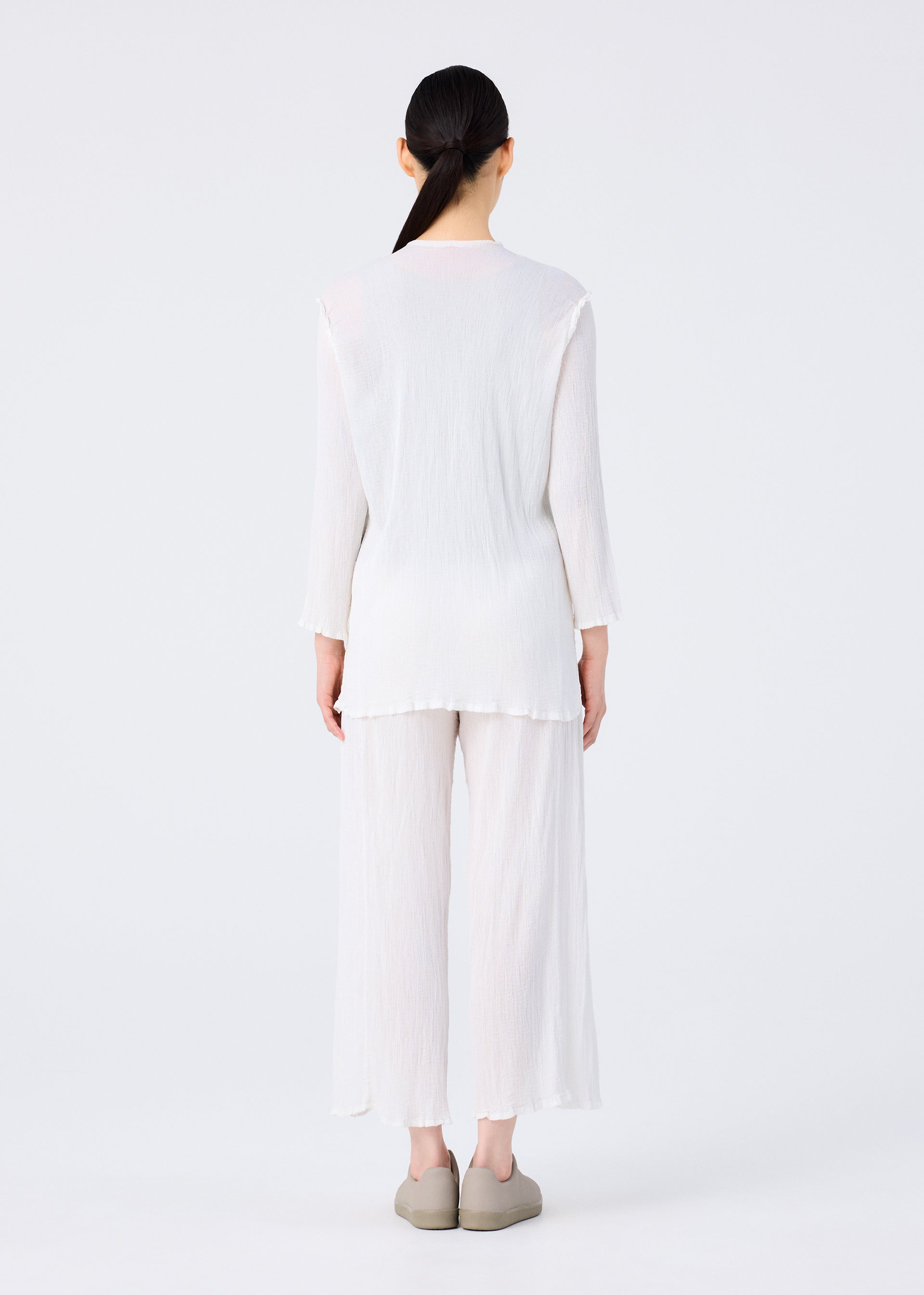 Zoomed view A person with long dark hair in a low ponytail faces away, wearing me ISSEY MIYAKE's SEE-THROUGH CREPE COLOR: a loose white long-sleeve top and matching see-through crepe pants, with light shoes, against a plain white background.