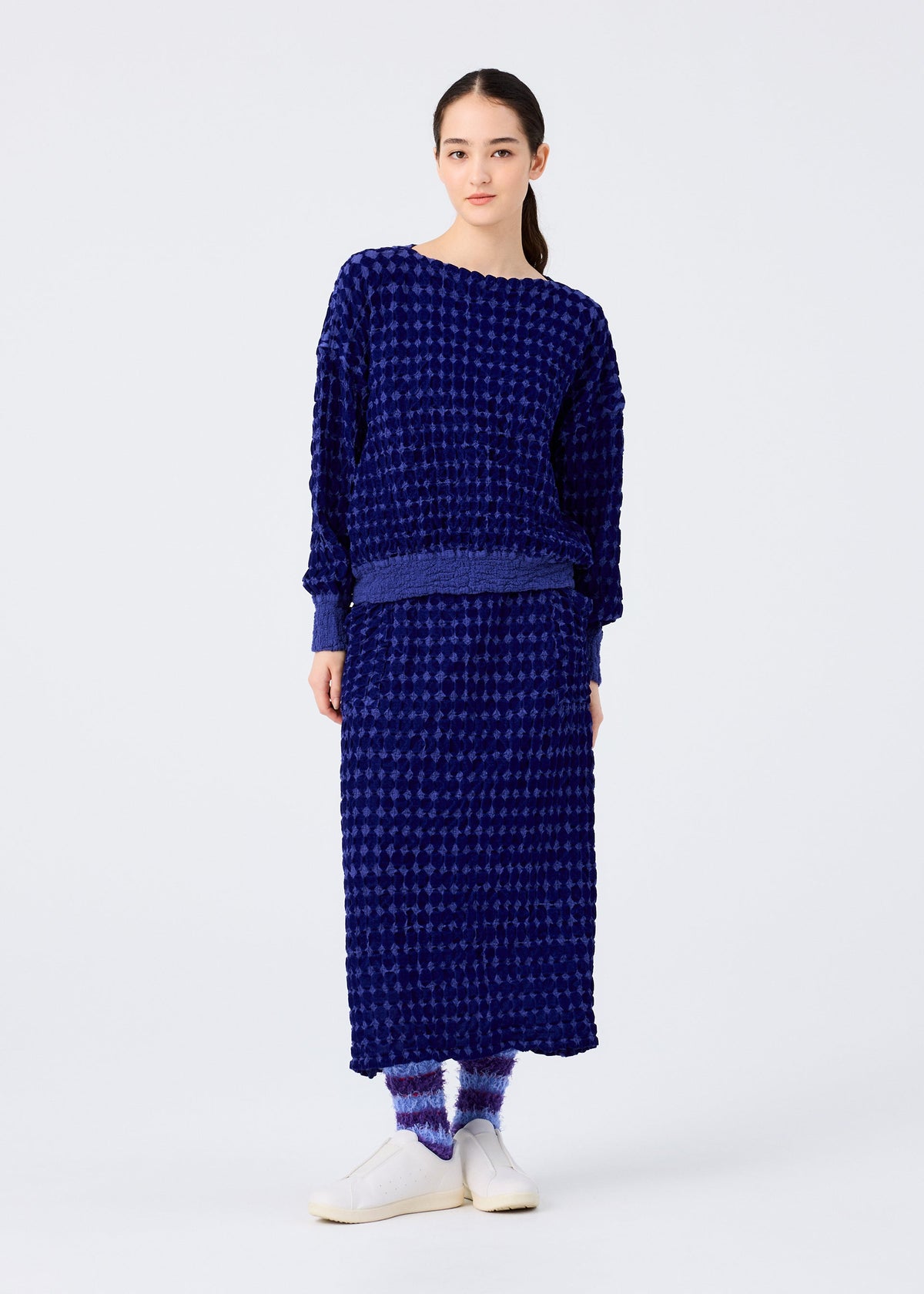 A woman with tied-back dark hair models the me ISSEY MIYAKE DIAMOND FLOCKY CAULIFLOWER: a textured dark blue long-sleeve top and matching cotton skirt with subtle flock printing, paired with blue-and-white socks and white slip-on shoes. view 5 of 6