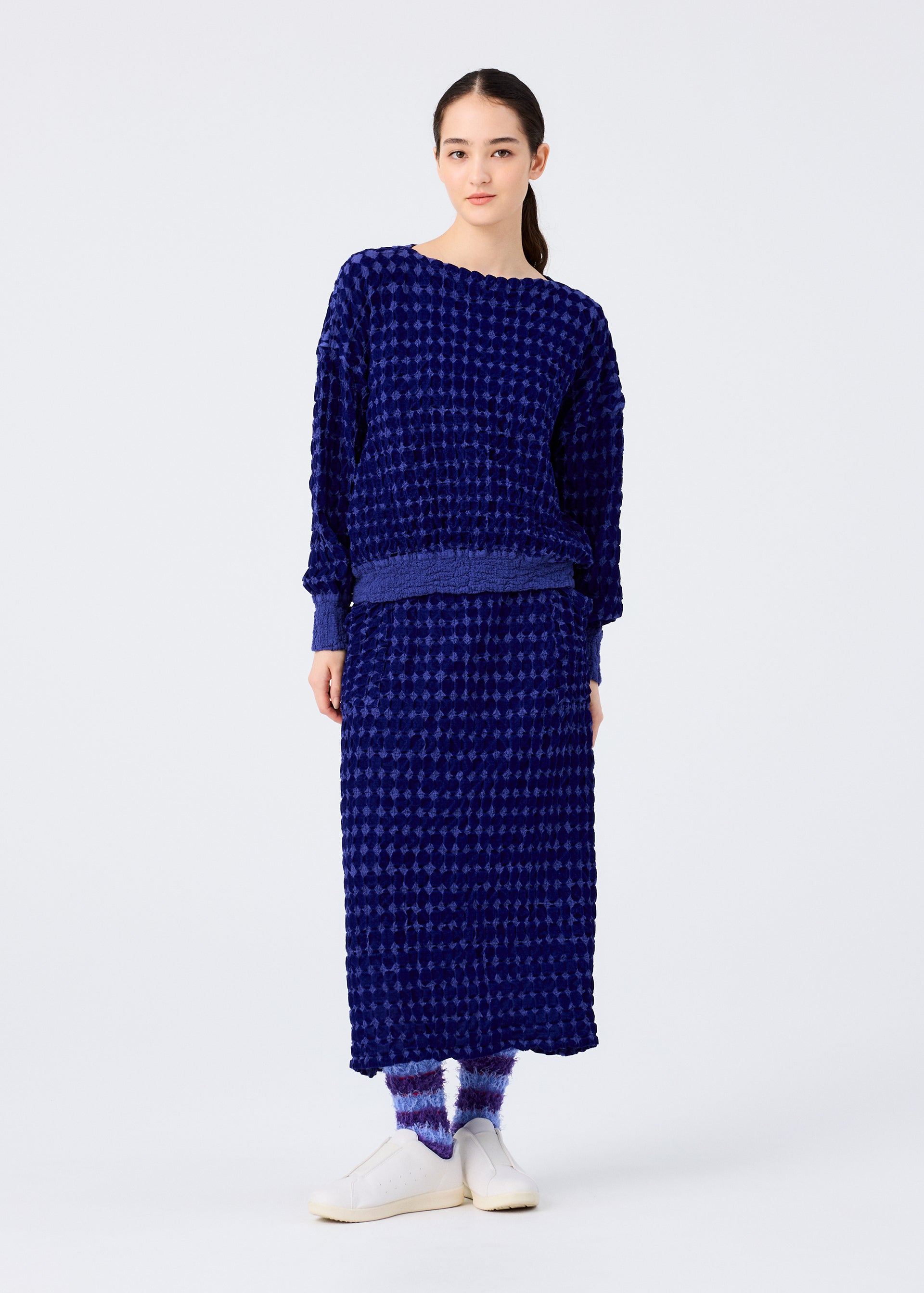Zoomed view A woman with tied-back dark hair models the me ISSEY MIYAKE DIAMOND FLOCKY CAULIFLOWER: a textured dark blue long-sleeve top and matching cotton skirt with subtle flock printing, paired with blue-and-white socks and white slip-on shoes.