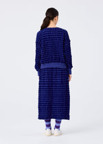 A person with long dark hair, seen from behind, models the me ISSEY MIYAKE DIAMOND FLOCKY CAULIFLOWER—textured dark blue sweater and matching diamond-patterned skirt—paired with purple socks and white shoes against a white background.