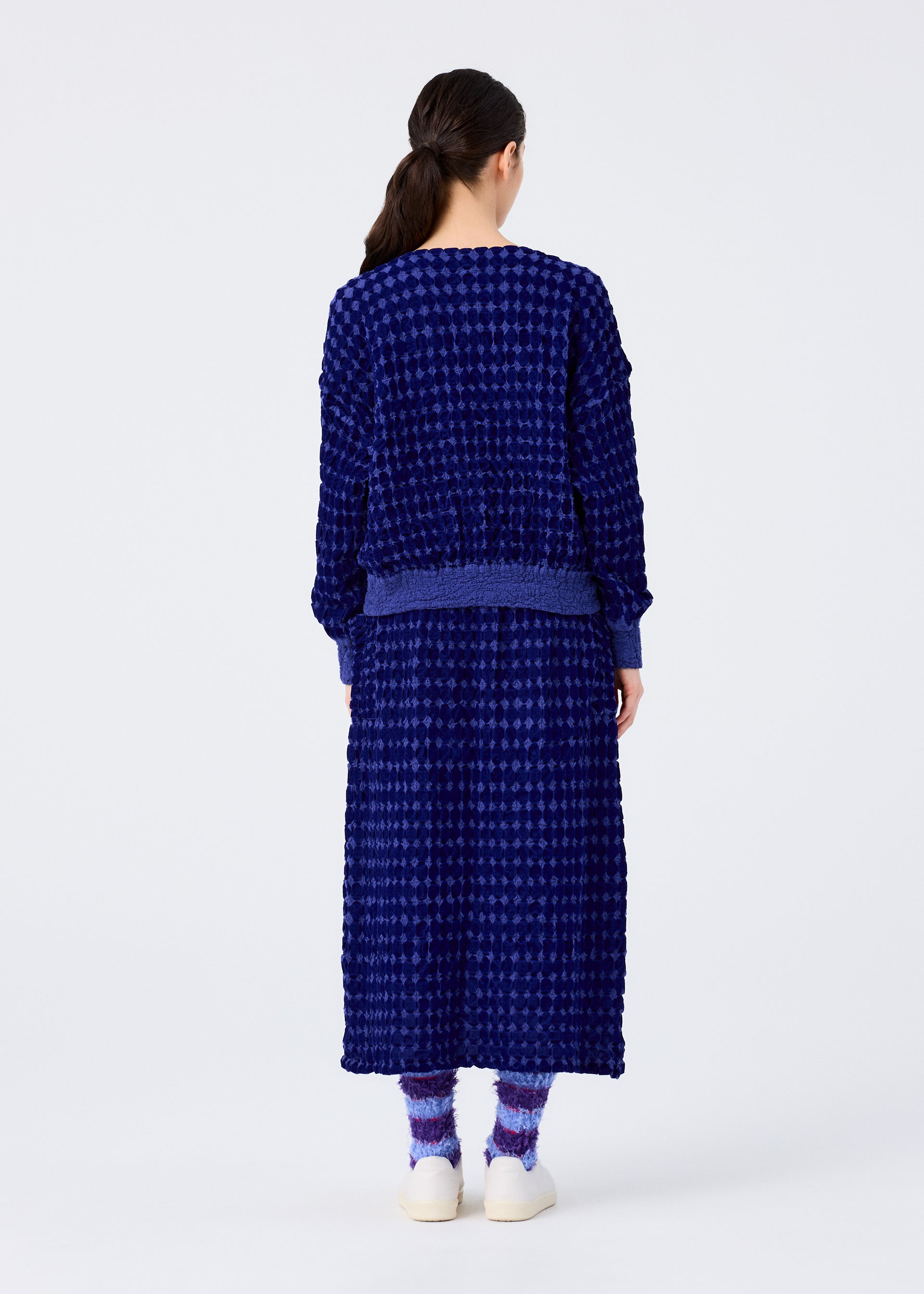 Zoomed view A person with long dark hair, seen from behind, models the me ISSEY MIYAKE DIAMOND FLOCKY CAULIFLOWER—textured dark blue sweater and matching diamond-patterned skirt—paired with purple socks and white shoes against a white background.