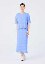A woman faces forward, smiling in the RIPPLE SEE-THROUGH CREPE by me ISSEY MIYAKE—a loose-fitting light blue short-sleeve top and matching pleated long skirt with subtle texture—plus white slip-on shoes against a plain light background.