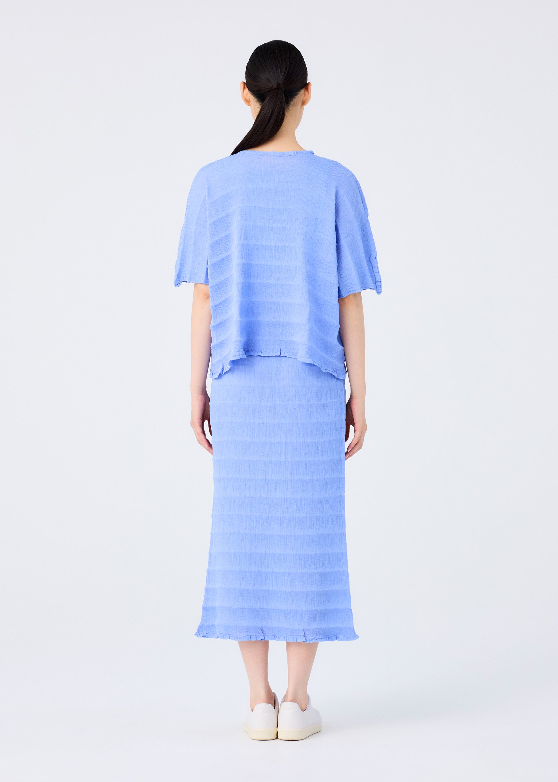 Zoomed view A woman with dark hair in a low ponytail, seen from behind, wears the me ISSEY MIYAKE RIPPLE SEE-THROUGH CREPE light blue top and matching midi skirt with textured stripes, paired with white shoes, against a plain white background.