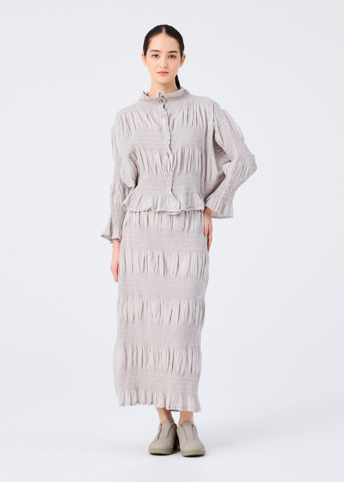 A woman faces forward wearing the SHRINK STRIPE light gray textured long-sleeve top and matching cotton blend skirt by me ISSEY MIYAKE, with her hair pulled back and gray slip-on shoes, posed against a plain white background. view 5 of 6