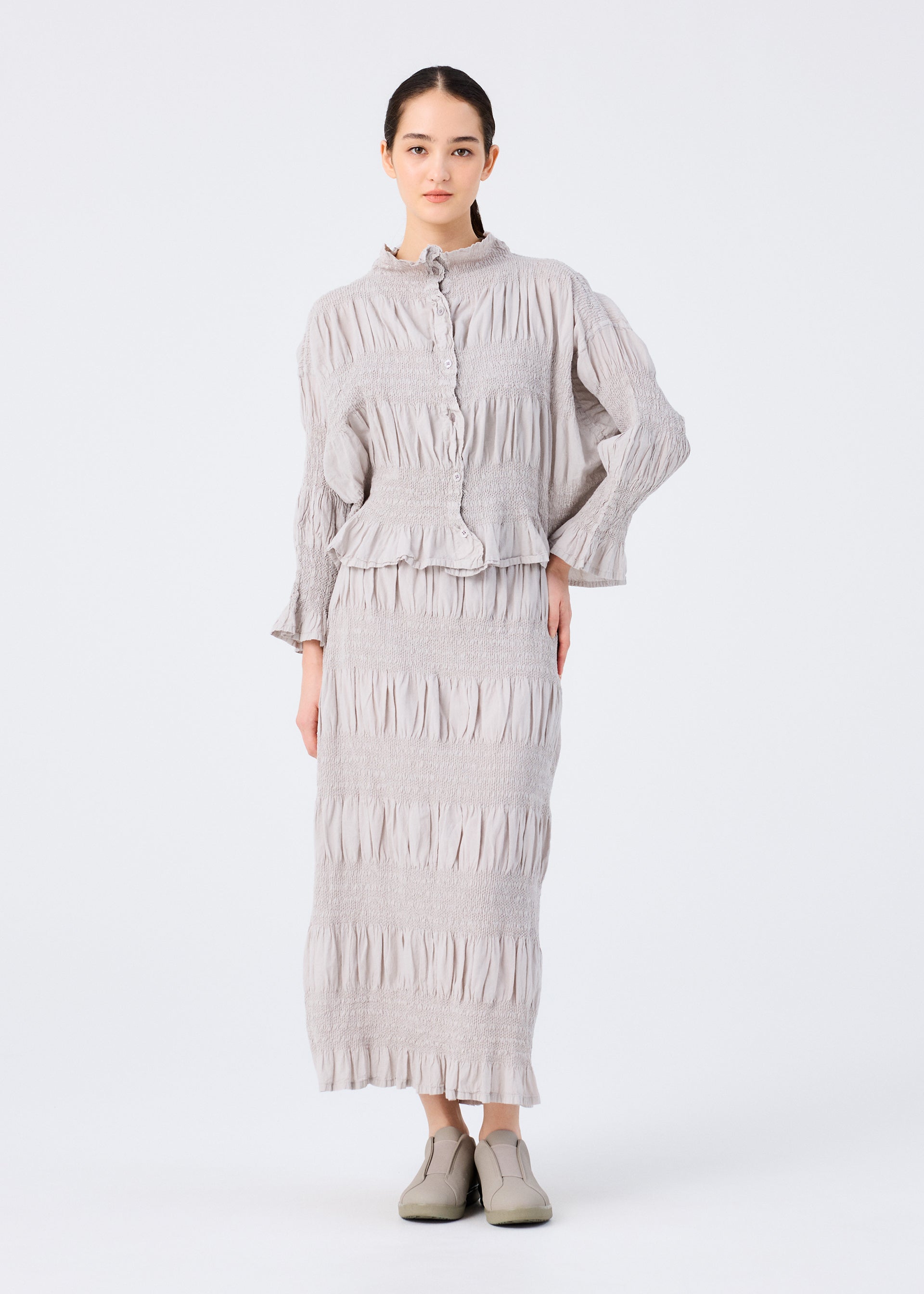 Zoomed view A woman faces forward wearing the SHRINK STRIPE light gray textured long-sleeve top and matching cotton blend skirt by me ISSEY MIYAKE, with her hair pulled back and gray slip-on shoes, posed against a plain white background.