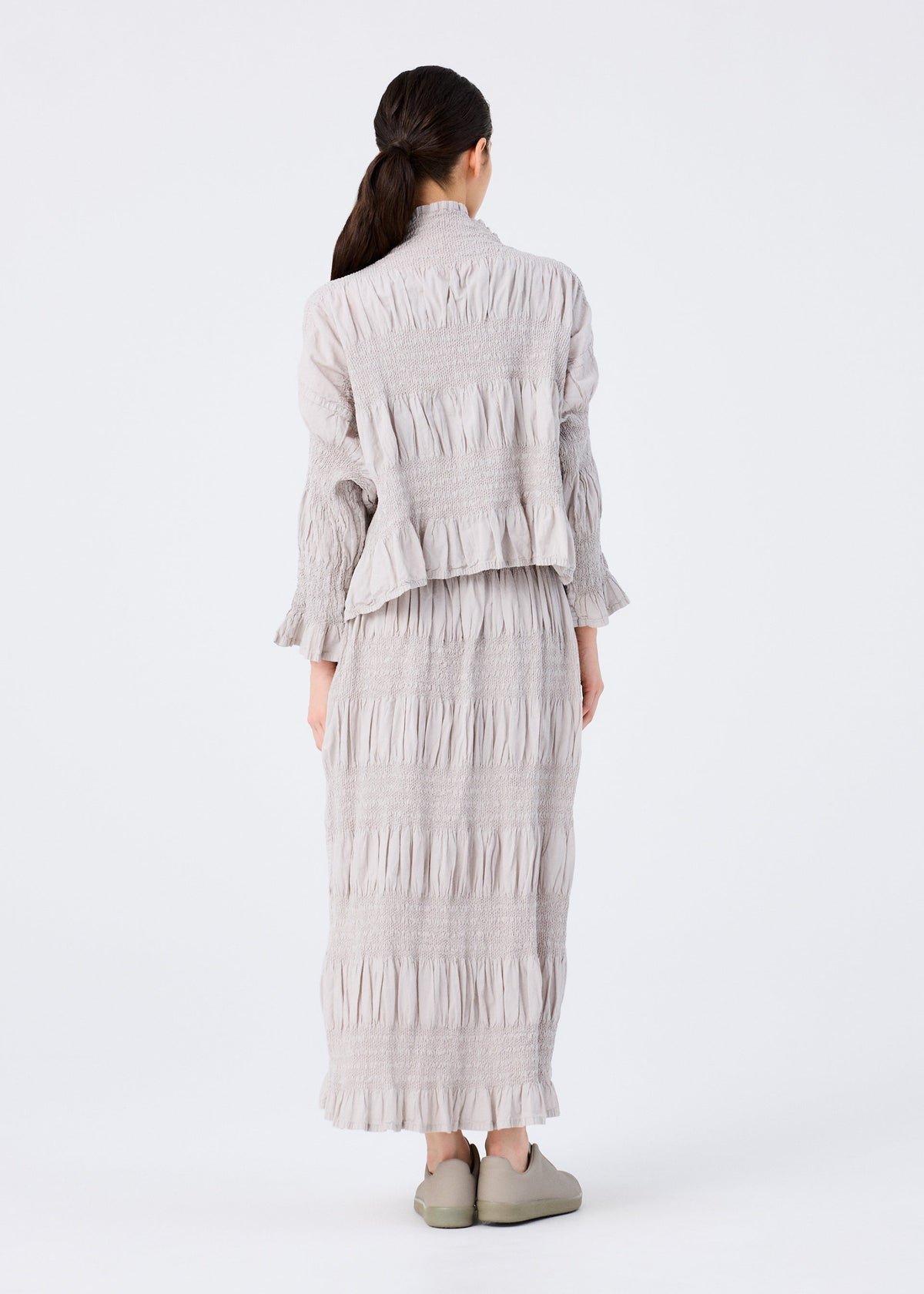 A person with long dark hair tied back faces away, wearing the SHRINK STRIPE long-sleeved top and matching skirt by me ISSEY MIYAKE in light gray with matching shoes, set against a plain white background. view 6 of 6