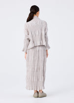 A person with long dark hair tied back faces away, wearing the SHRINK STRIPE long-sleeved top and matching skirt by me ISSEY MIYAKE in light gray with matching shoes, set against a plain white background.