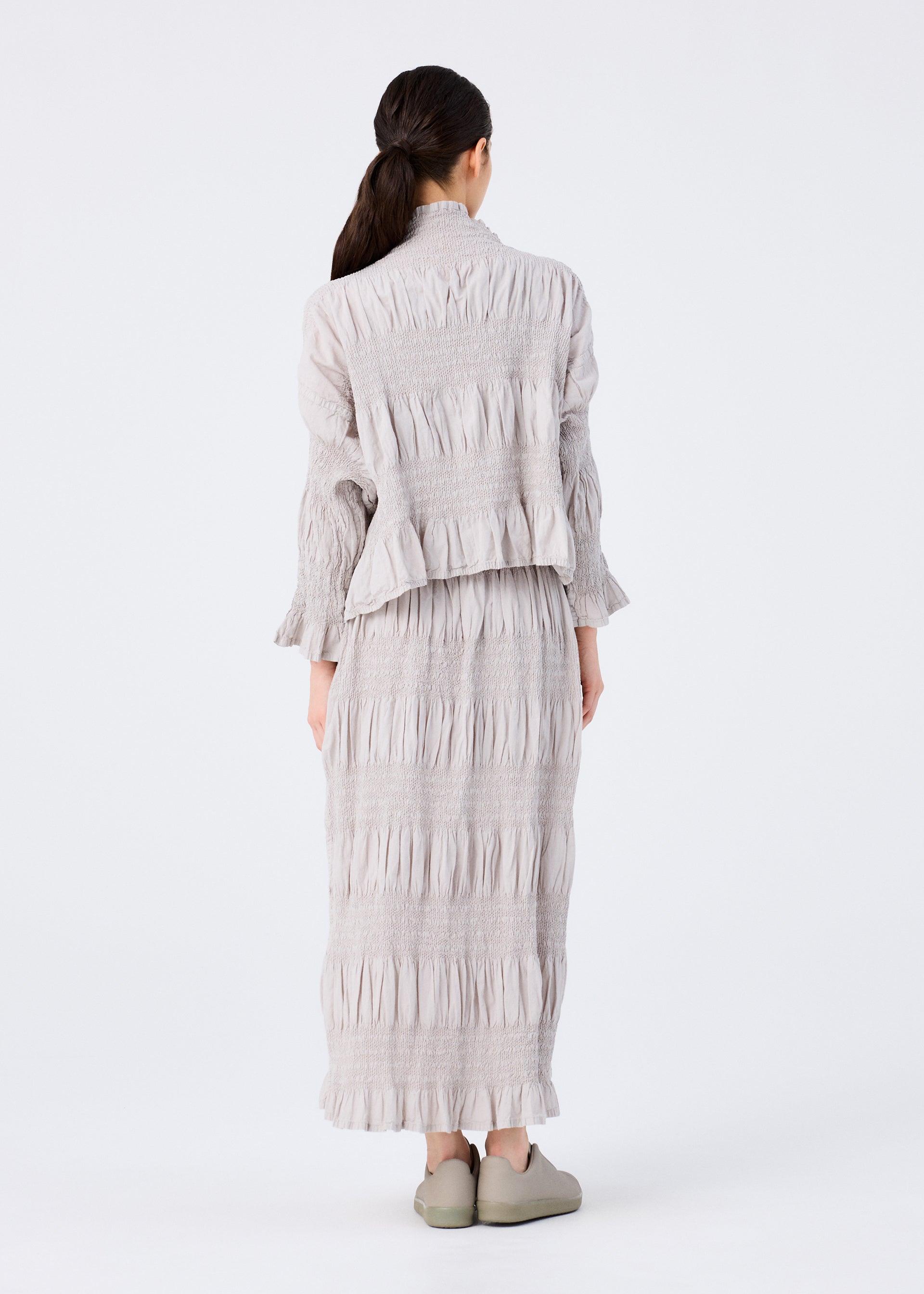 Zoomed view A person with long dark hair tied back faces away, wearing the SHRINK STRIPE long-sleeved top and matching skirt by me ISSEY MIYAKE in light gray with matching shoes, set against a plain white background.