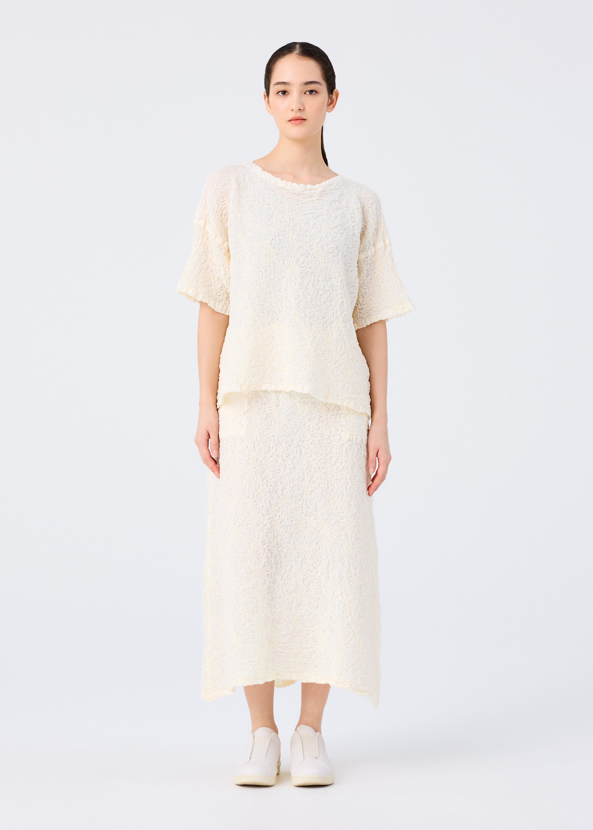 Zoomed view A person stands facing forward against a white background, wearing the SURPRISE BOX PRINT CAULIFLOWER set by me ISSEY MIYAKE: a loose, textured cream top with short sleeves and a matching cotton skirt. Their dark hair is pulled back; they wear white shoes.