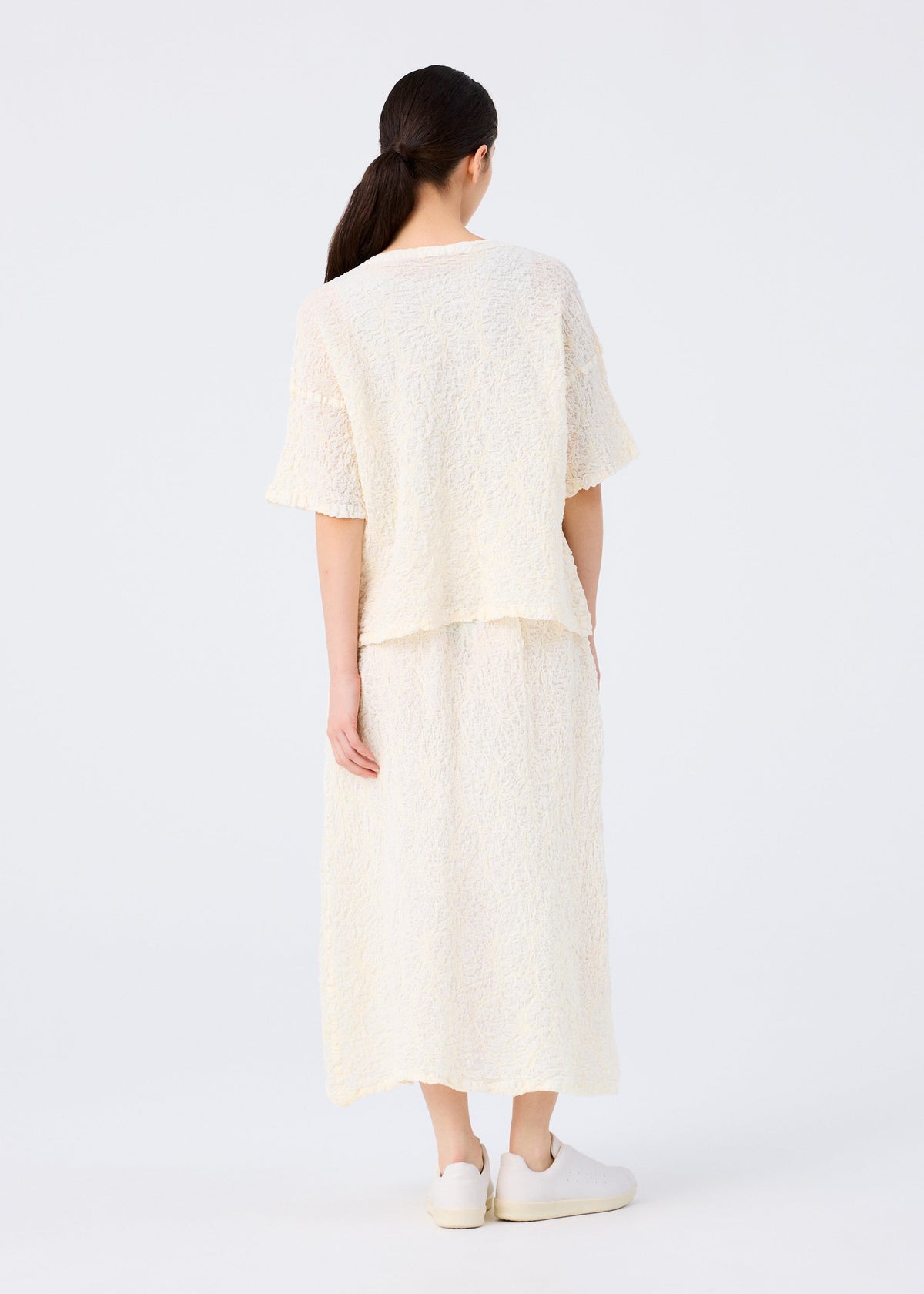 A woman with long dark hair in a low ponytail, wearing the SURPRISE BOX PRINT CAULIFLOWER set from me ISSEY MIYAKE and white sneakers, stands facing away against a plain light background. view 6 of 6