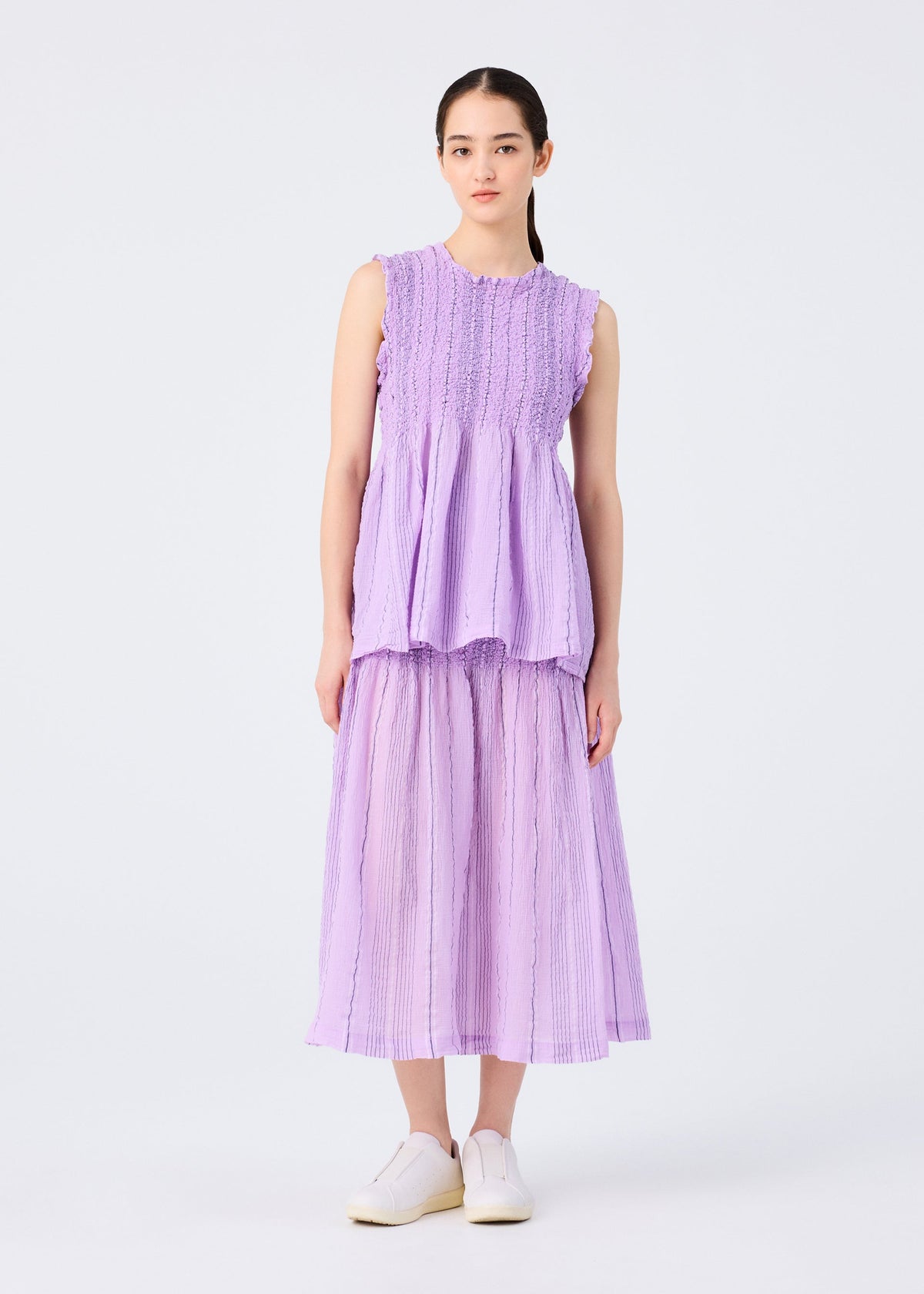 A woman stands against a plain white background wearing the SHIRT STRIPE CAULIFLOWER by me ISSEY MIYAKE—a sleeveless, textured lavender top with a matching pleated cotton skirt—paired with white slip-on shoes. view 5 of 6