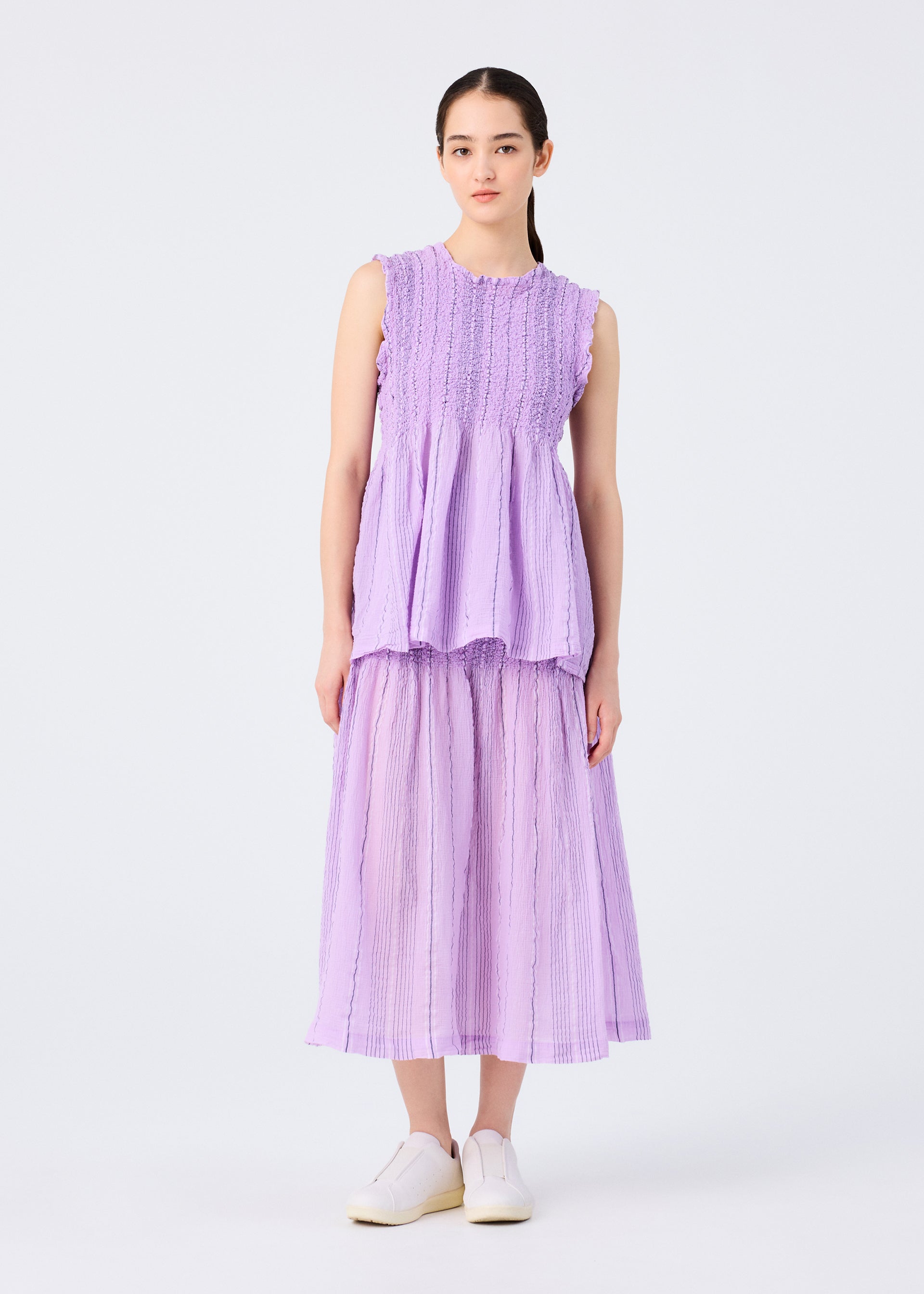 Zoomed view A woman stands against a plain white background wearing the SHIRT STRIPE CAULIFLOWER by me ISSEY MIYAKE—a sleeveless, textured lavender top with a matching pleated cotton skirt—paired with white slip-on shoes.