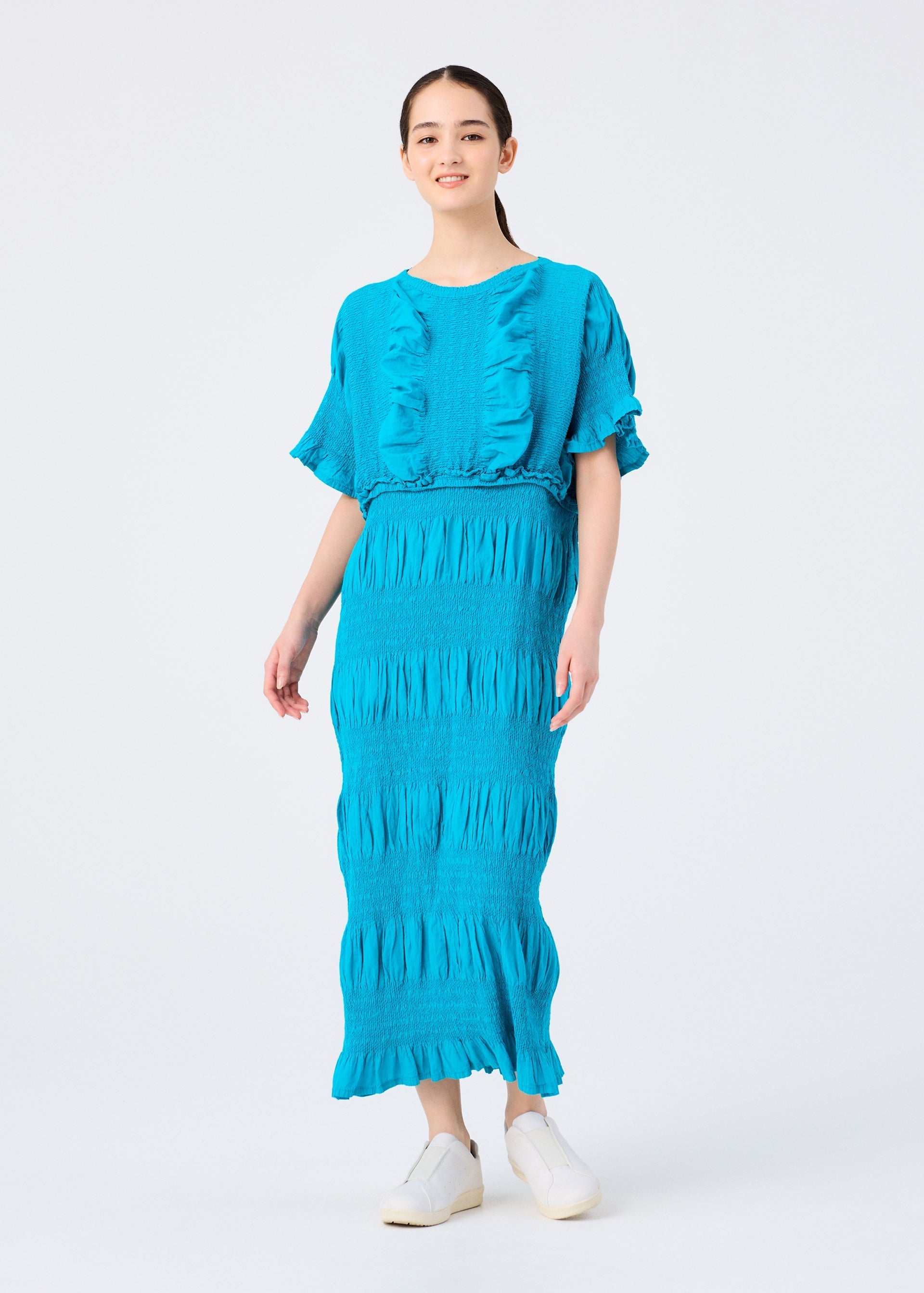 Zoomed view A woman with dark hair tied back smiles in a bright blue me ISSEY MIYAKE SHRINK STRIPE textured cotton-blend dress with short sleeves and matching long skirt, against a white background. She pairs it with white slip-on shoes.