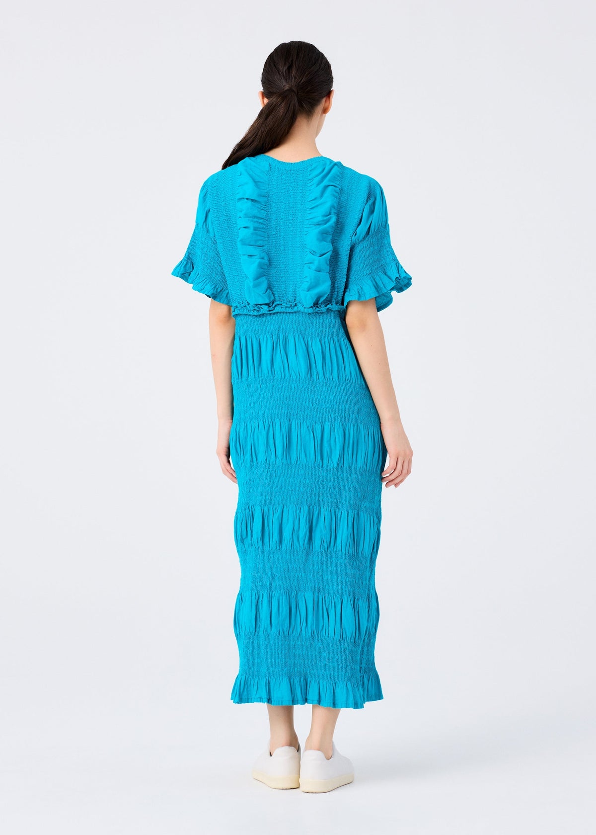 A woman with long dark hair faces away from the camera, wearing the me ISSEY MIYAKE SHRINK STRIPE cotton blend midi dress in bright blue with ruffle details and product-dyeing, paired with white slip-on shoes against a light background. view 6 of 6