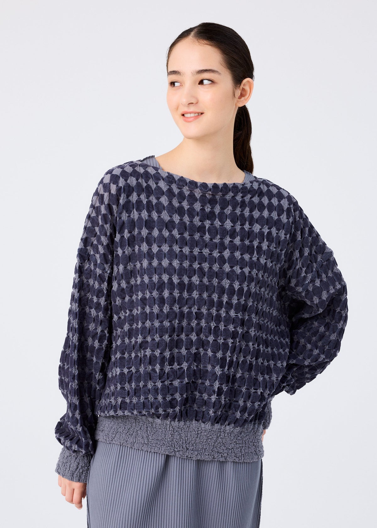 A woman with long dark hair in a ponytail wears the DIAMOND FLOCKY CAULIFLOWER top by me ISSEY MIYAKE and a pleated gray skirt, standing before a plain light background and smiling slightly while looking to the side. view 4 of 6