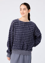 A woman with long dark hair in a ponytail wears the DIAMOND FLOCKY CAULIFLOWER top by me ISSEY MIYAKE and a pleated gray skirt, standing before a plain light background and smiling slightly while looking to the side.