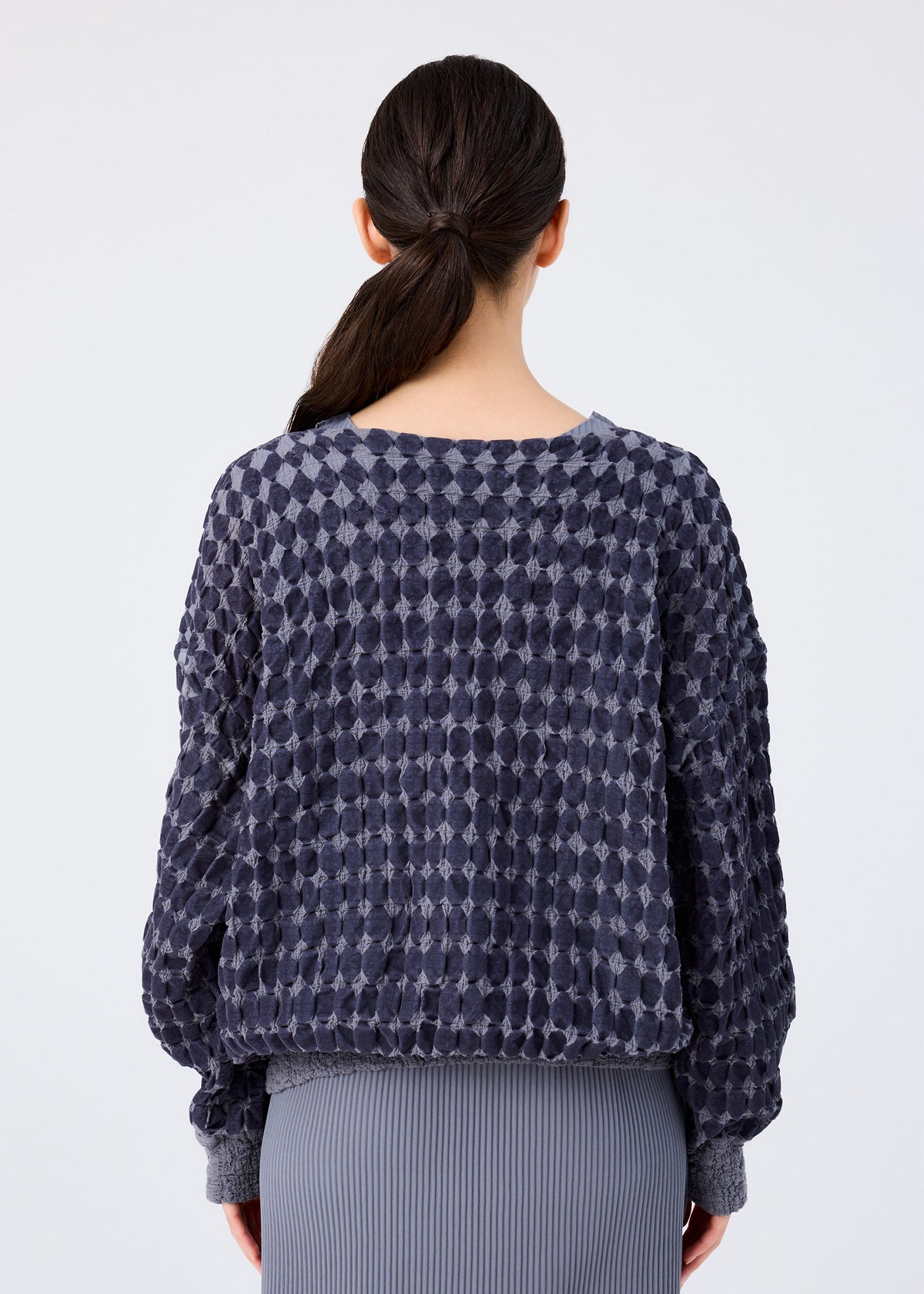 A woman with dark hair in a low ponytail, seen from the back, wears the me ISSEY MIYAKE DIAMOND FLOCKY CAULIFLOWER long-sleeved top with diamond-patterned flock print in navy and a gray pleated skirt. view 5 of 6