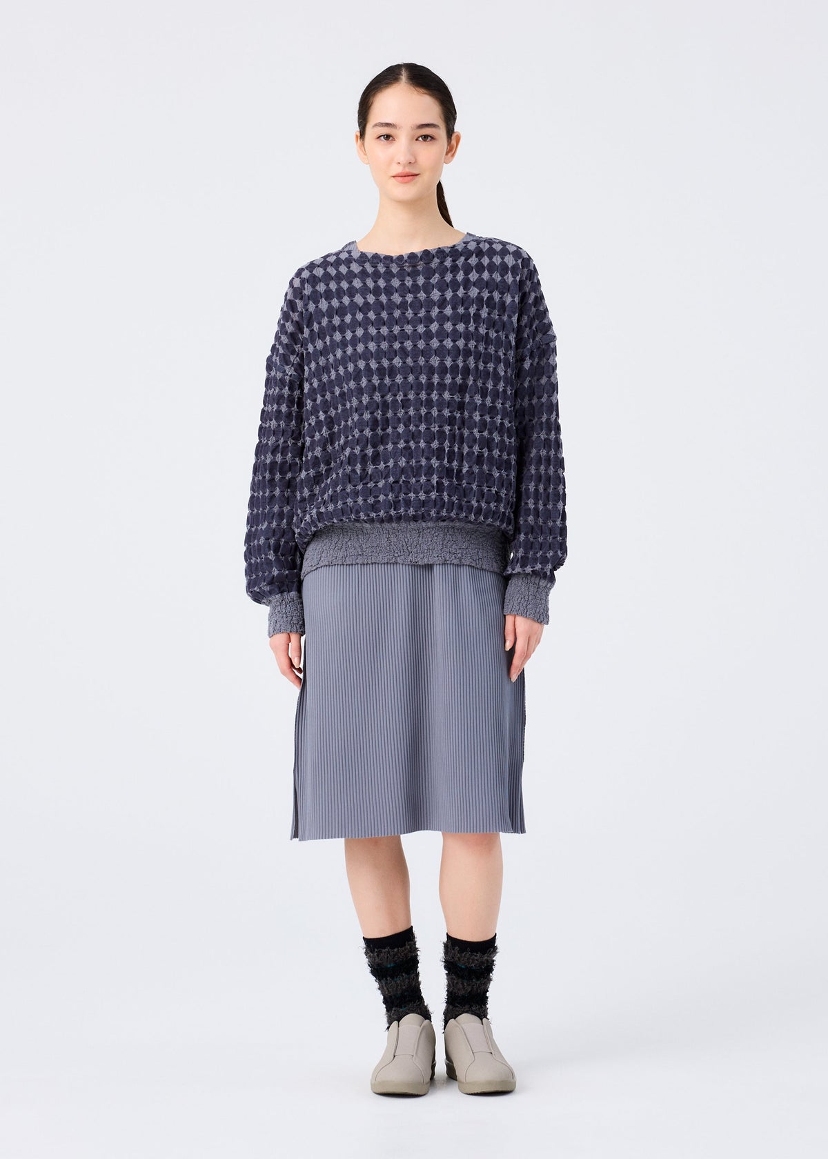 A woman stands against a white background, wearing a loose, long-sleeved top in dark blue DIAMOND FLOCKY CAULIFLOWER by me ISSEY MIYAKE, a gray knee-length pleated skirt, black socks, and beige slip-on shoes. She faces forward neutrally. view 6 of 6