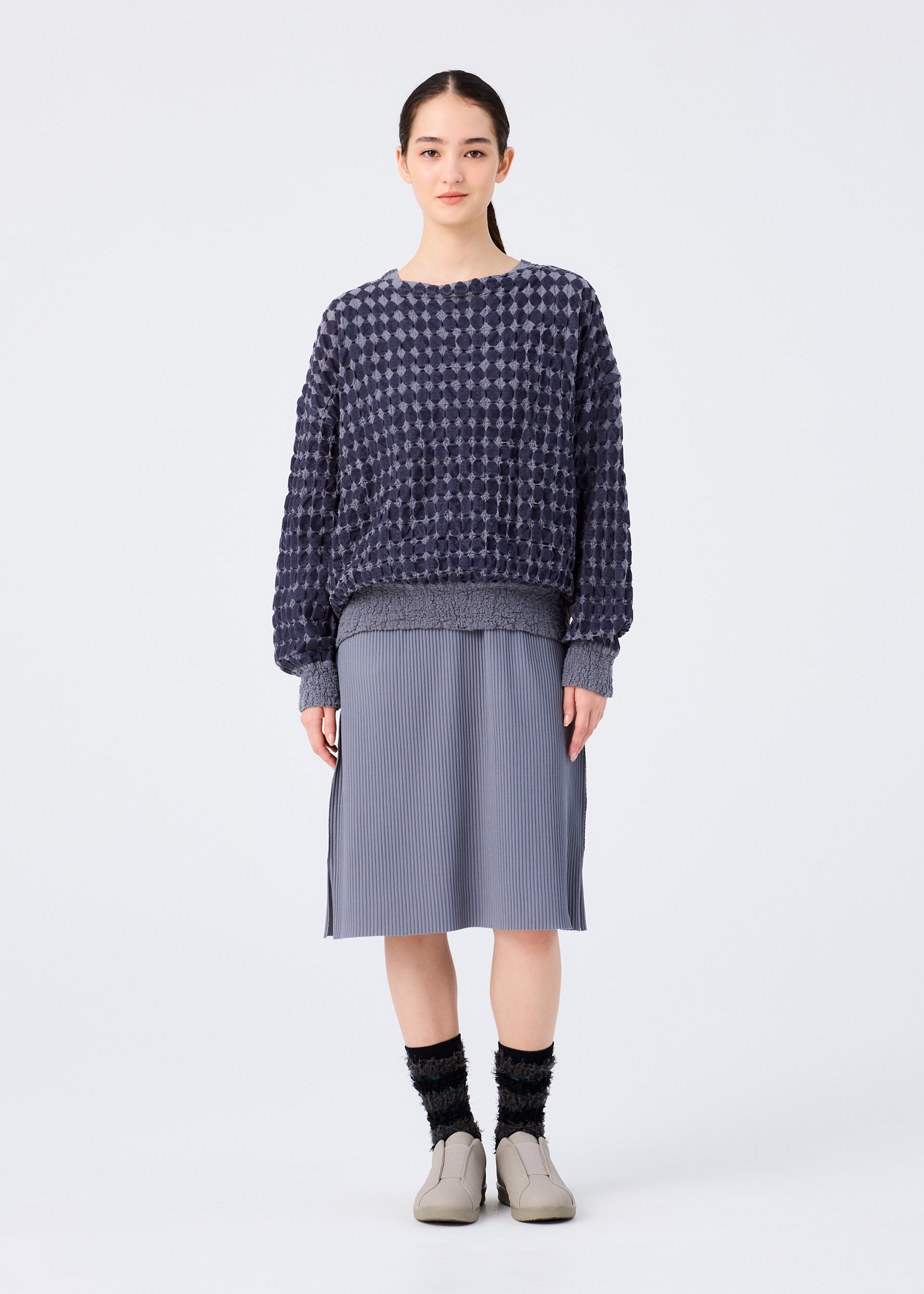 Zoomed view A woman stands against a white background, wearing a loose, long-sleeved top in dark blue DIAMOND FLOCKY CAULIFLOWER by me ISSEY MIYAKE, a gray knee-length pleated skirt, black socks, and beige slip-on shoes. She faces forward neutrally.