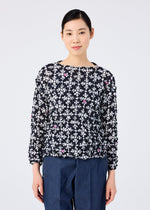 A woman with dark hair pulled back wears the SUIT MERINGUE TOP by me ISSEY MIYAKE, a round-neck long-sleeved top with a white and pink floral pattern, paired with blue trousers against a plain white background.