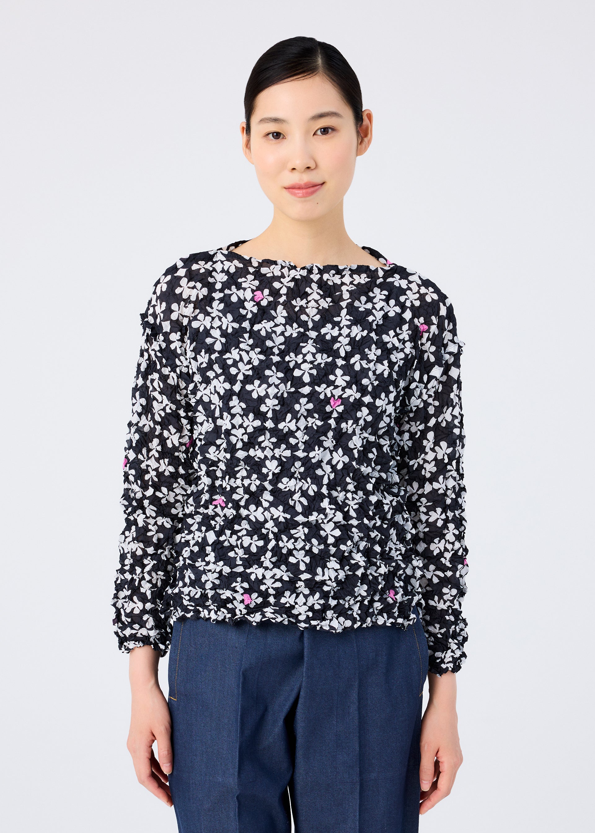 Zoomed view A woman with dark hair pulled back wears the SUIT MERINGUE TOP by me ISSEY MIYAKE, a round-neck long-sleeved top with a white and pink floral pattern, paired with blue trousers against a plain white background.
