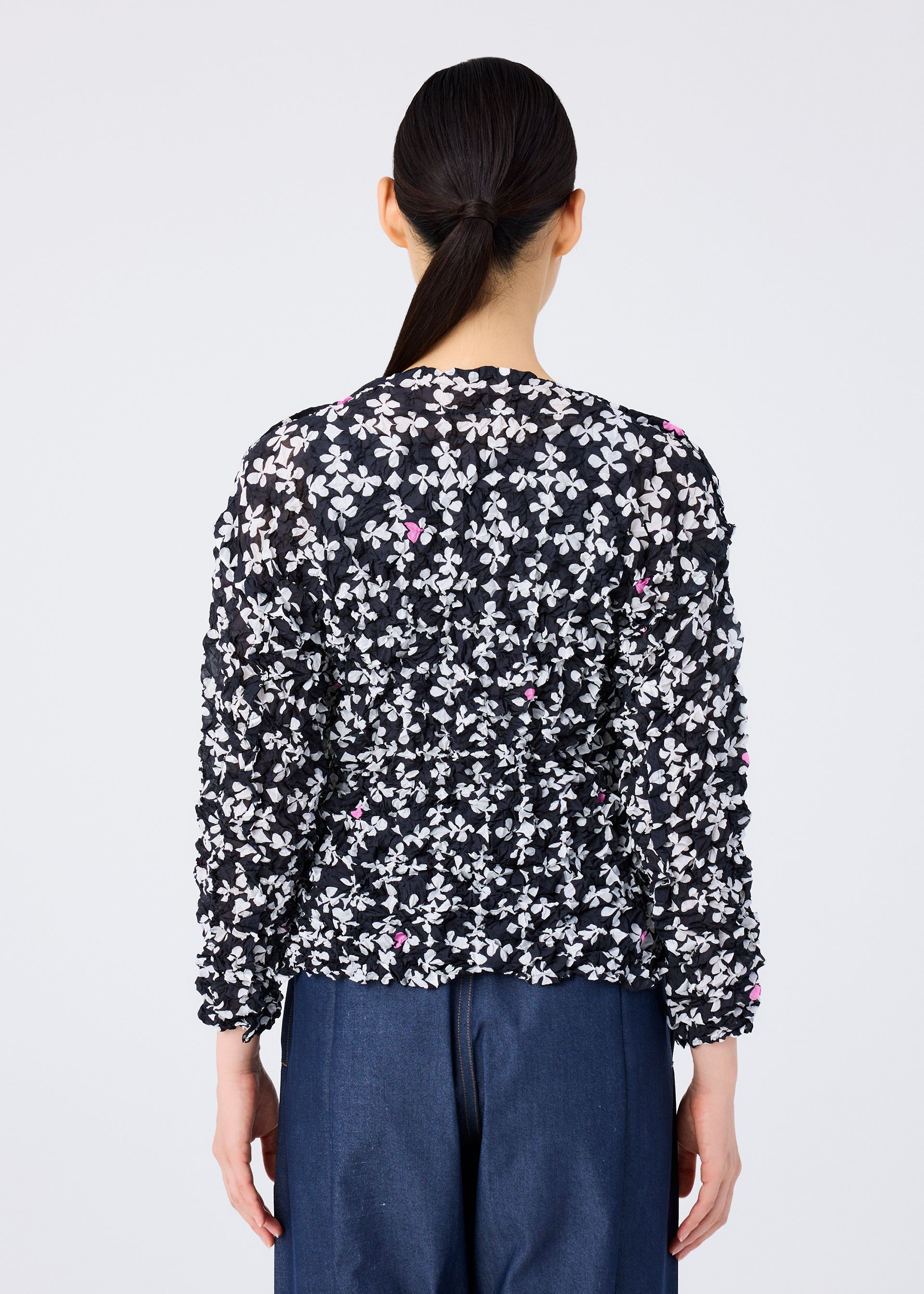 Zoomed view A person with long dark hair in a low ponytail is seen from behind wearing the SUIT MERINGUE TOP by me ISSEY MIYAKE, which features white and pink floral patterns, paired with blue pants against a plain light background.