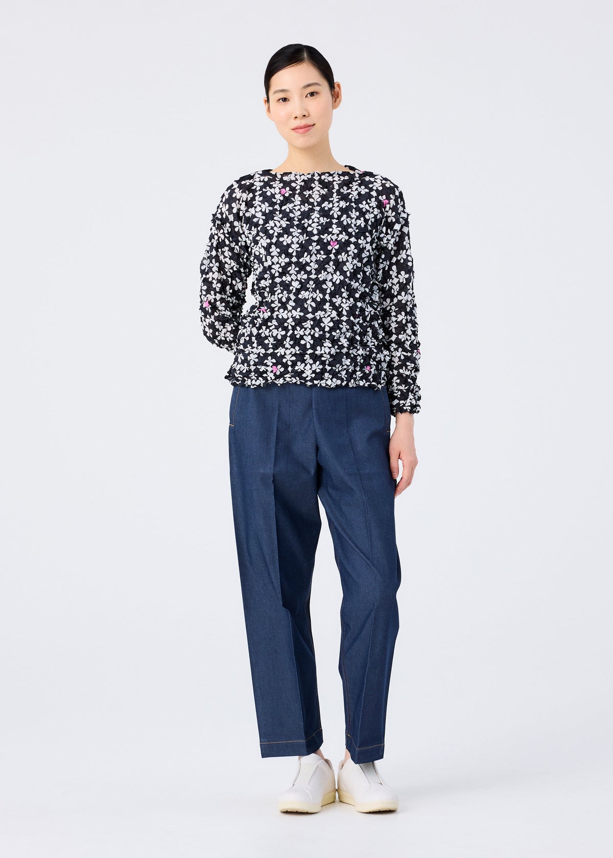 A woman stands against a white background, wearing the SUIT MERINGUE TOP by me ISSEY MIYAKE—a black round-neck, long-sleeve top with a white floral pattern. She pairs it with loose blue pants, white shoes, and has her hair tied back with a slight smile. view 8 of 8
