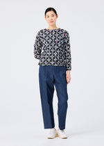 A woman stands against a white background, wearing the SUIT MERINGUE TOP by me ISSEY MIYAKE—a black round-neck, long-sleeve top with a white floral pattern. She pairs it with loose blue pants, white shoes, and has her hair tied back with a slight smile.