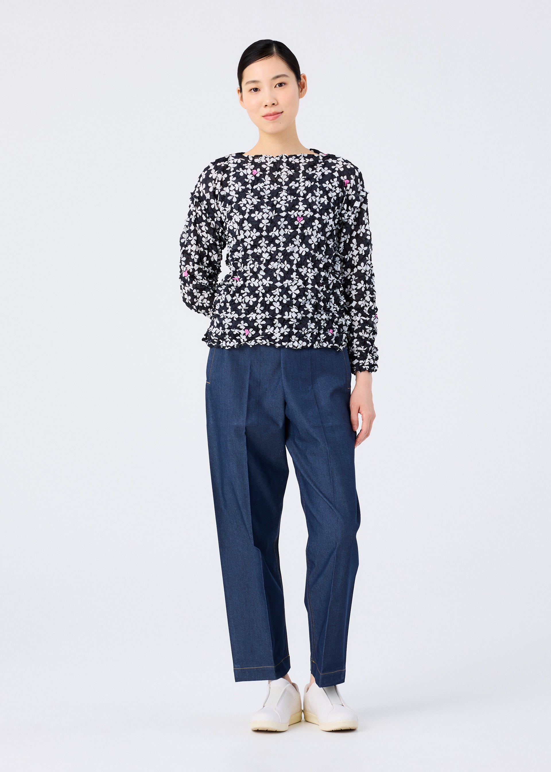 Zoomed view A woman stands against a white background, wearing the SUIT MERINGUE TOP by me ISSEY MIYAKE—a black round-neck, long-sleeve top with a white floral pattern. She pairs it with loose blue pants, white shoes, and has her hair tied back with a slight smile.
