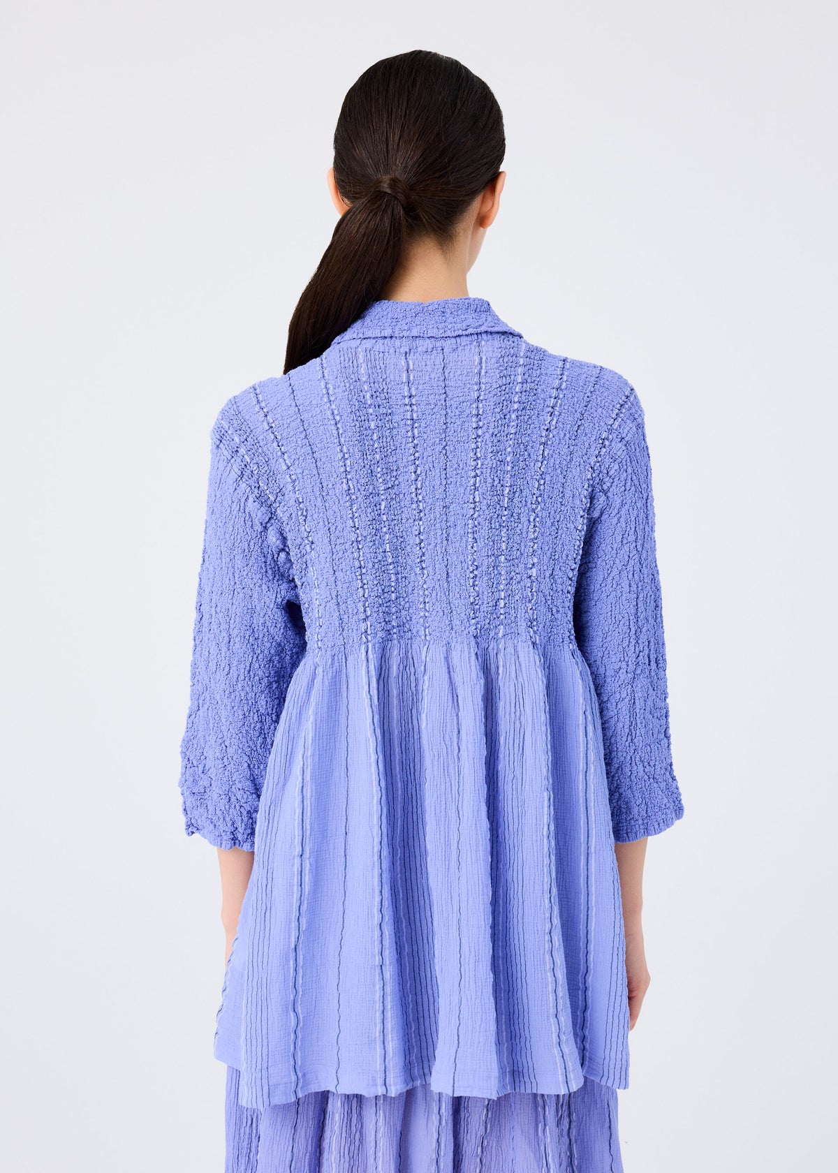 A woman with long dark hair in a low ponytail is seen from the back, wearing the me ISSEY MIYAKE SHIRT STRIPE CAULIFLOWER—a textured, light purple, long-sleeved shirt with a flared hem and vertical seams—against a plain white background. view 6 of 7