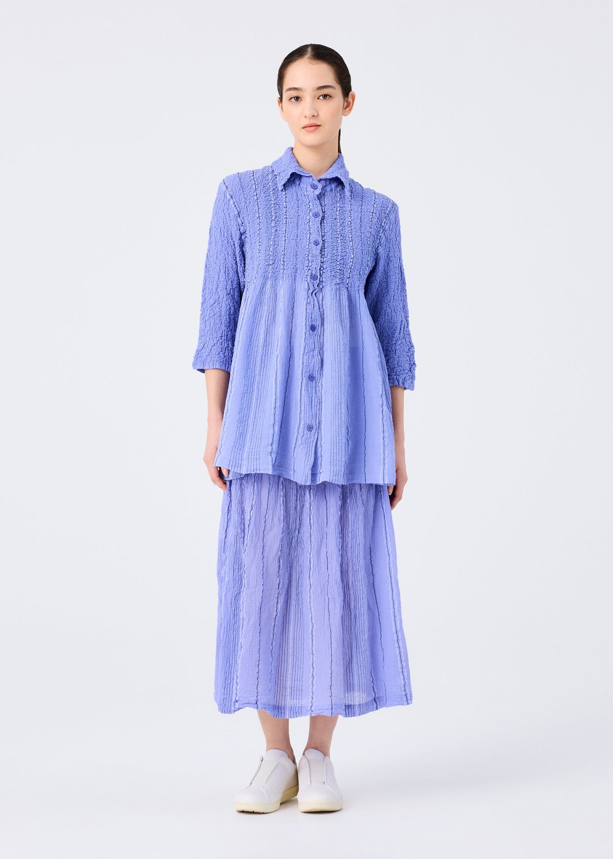 A woman stands against a white background wearing the SHIRT STRIPE CAULIFLOWER by me ISSEY MIYAKE—a loose, textured light blue blouse with subtle stripes and three-quarter sleeves—paired with a matching pleated skirt and white slip-ons. view 7 of 7