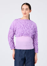 A woman with straight dark hair pulled back stands against a plain background wearing the MERINGUE TOP by me ISSEY MIYAKE, a crushed pleat, long-sleeved light purple top with a crinkled pattern, paired with dark blue pants.