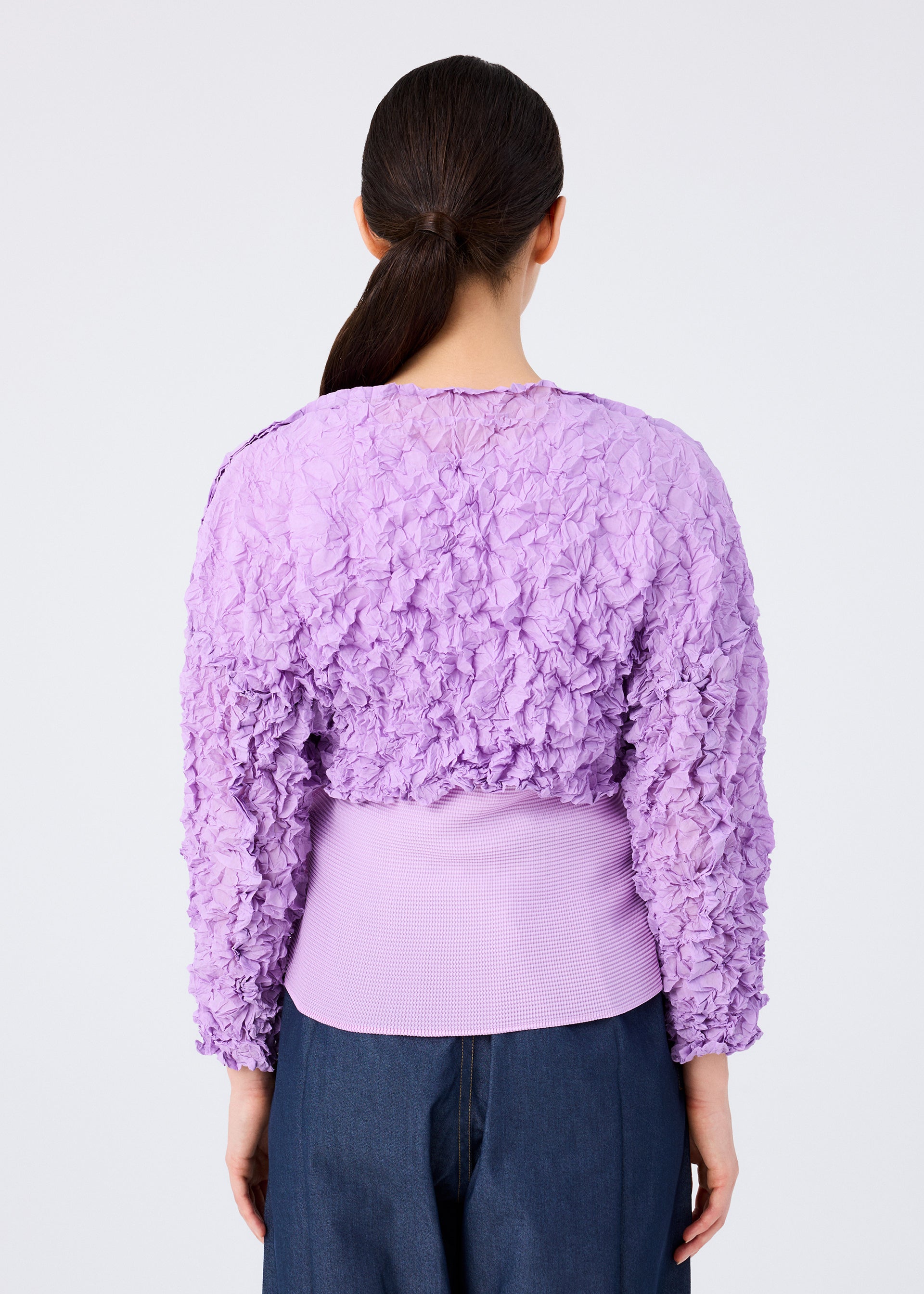 Zoomed view A woman with long dark hair in a low ponytail is seen from behind, wearing the me ISSEY MIYAKE MERINGUE TOP— a lavender crushed pleat, long-sleeved cropped top—paired with dark blue pants against a plain white background.