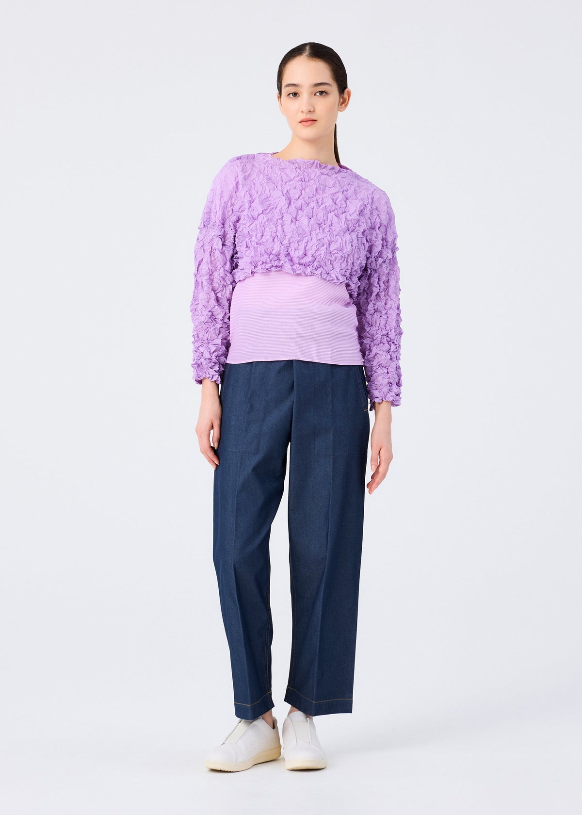 A woman wears the MERINGUE TOP by me ISSEY MIYAKE, a long-sleeved crushed pleat crop top, paired with a purple ribbed tank, dark denim wide-leg pants, and white sneakers. view 9 of 9
