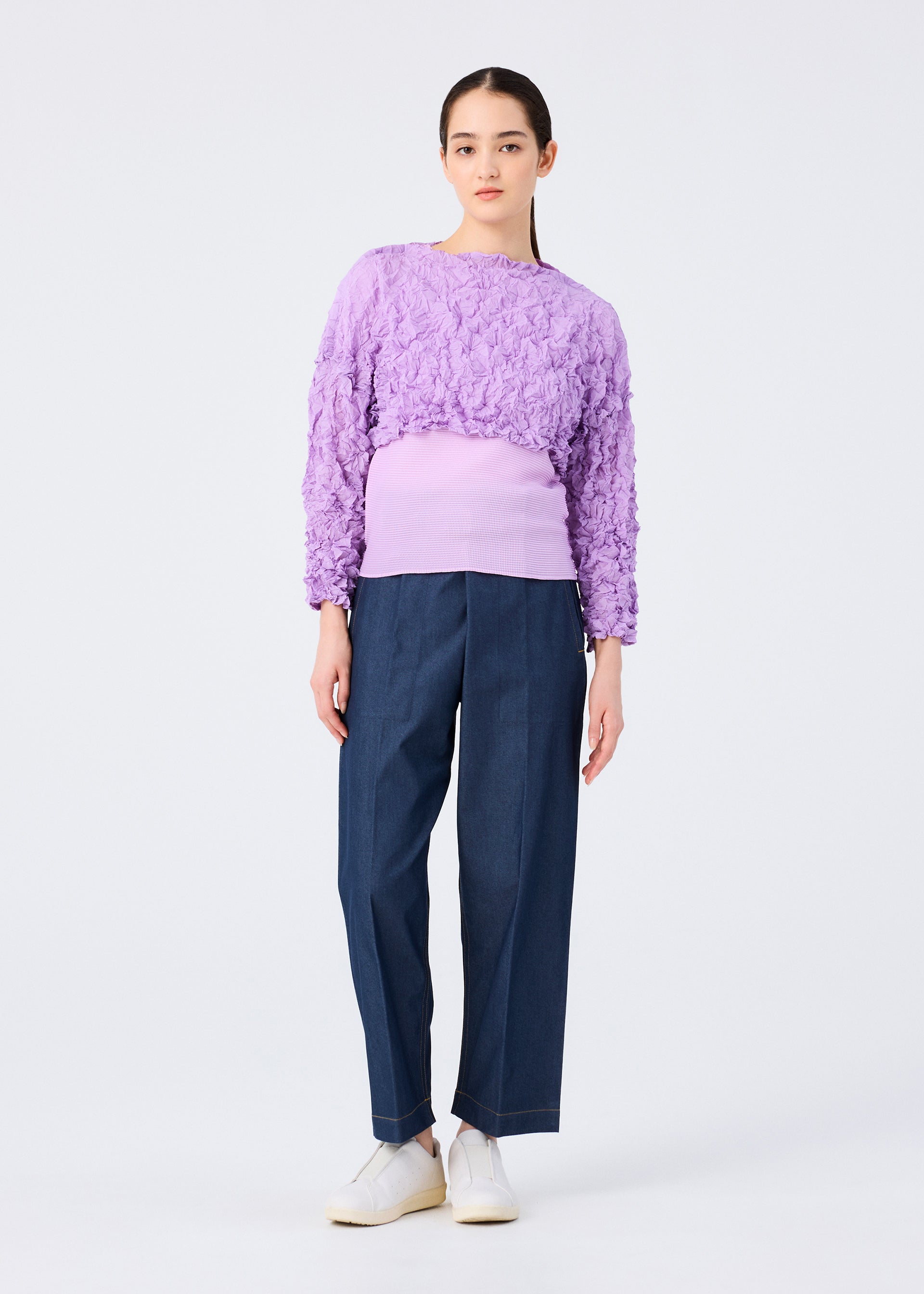 Zoomed view A woman wears the MERINGUE TOP by me ISSEY MIYAKE, a long-sleeved crushed pleat crop top, paired with a purple ribbed tank, dark denim wide-leg pants, and white sneakers.