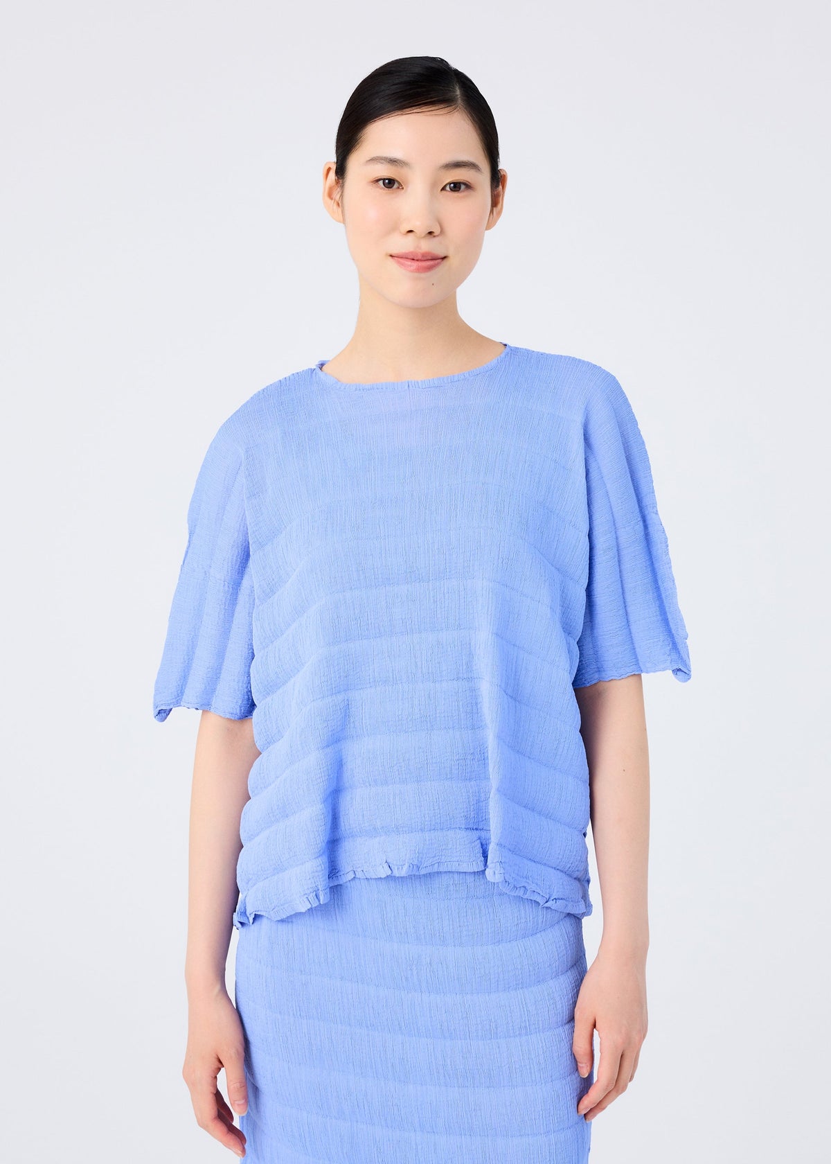 A person with dark hair tied back wears the me ISSEY MIYAKE RIPPLE SEE-THROUGH CREPE light blue, short-sleeve top and matching pleated skirt, standing against a plain white background. view 5 of 7