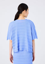 A person with long dark hair in a low ponytail wears the me ISSEY MIYAKE RIPPLE SEE-THROUGH CREPE light blue top and matching skirt, shown from the back against a plain light background.