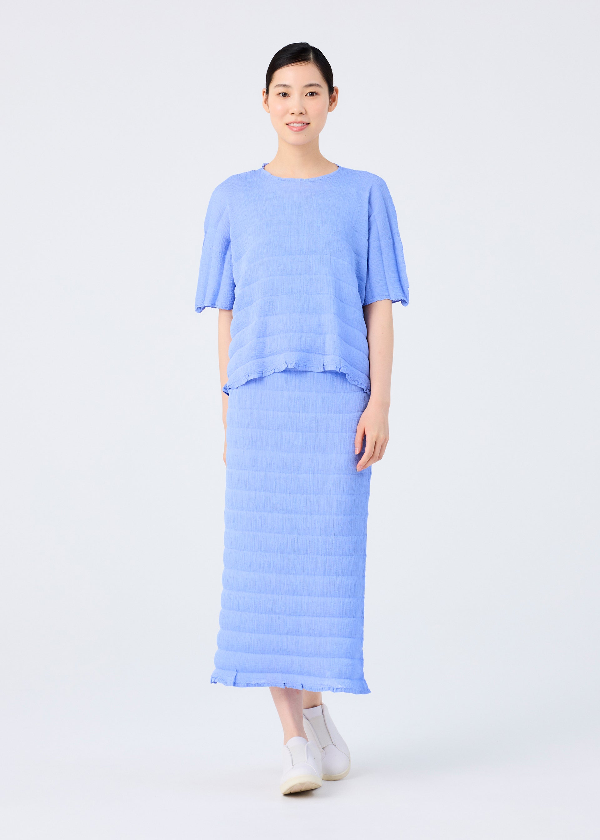 Zoomed view A person stands facing forward, wearing the me ISSEY MIYAKE RIPPLE SEE-THROUGH CREPE light blue textured short-sleeve top and matching long skirt with white shoes, set against a plain light background.