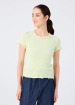 A woman with long dark hair in a low ponytail wears the me ISSEY MIYAKE CAULIFLOWER COLOR light yellow round-neck short-sleeved top and blue pants, standing and smiling slightly against a plain light background.